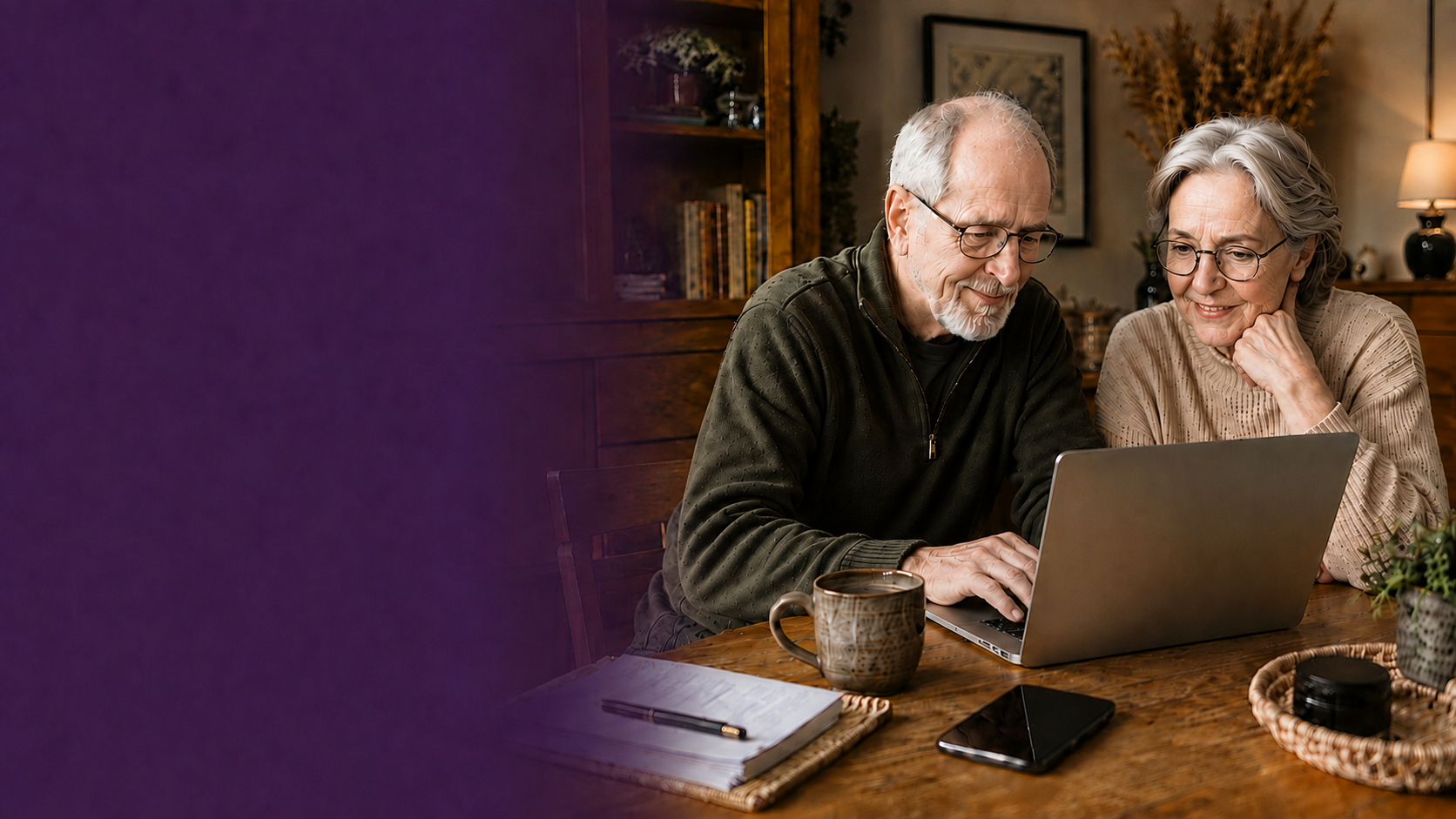 An older man and woman at their dining room table, both wearing glasses and smiling as they use a laptop together.